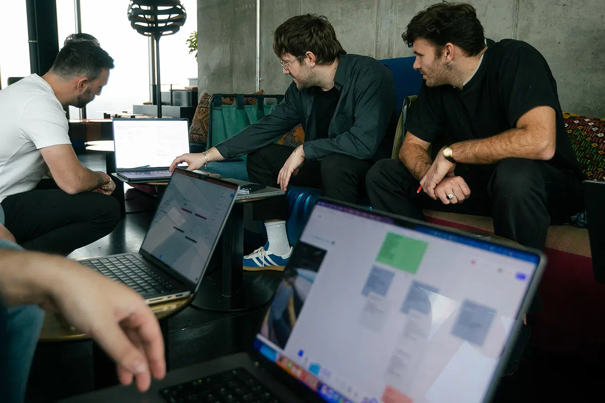 Three colleagues reviewing laptop screens together on a couch, one pointing at a document on his laptop while others look on.
