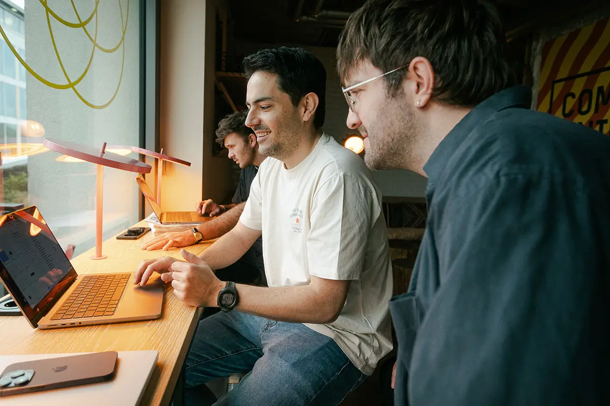 Man in a white t-shirt smiling while typing on a laptop at a wooden desk, with two other colleagues working on laptops in the background near a large window.
