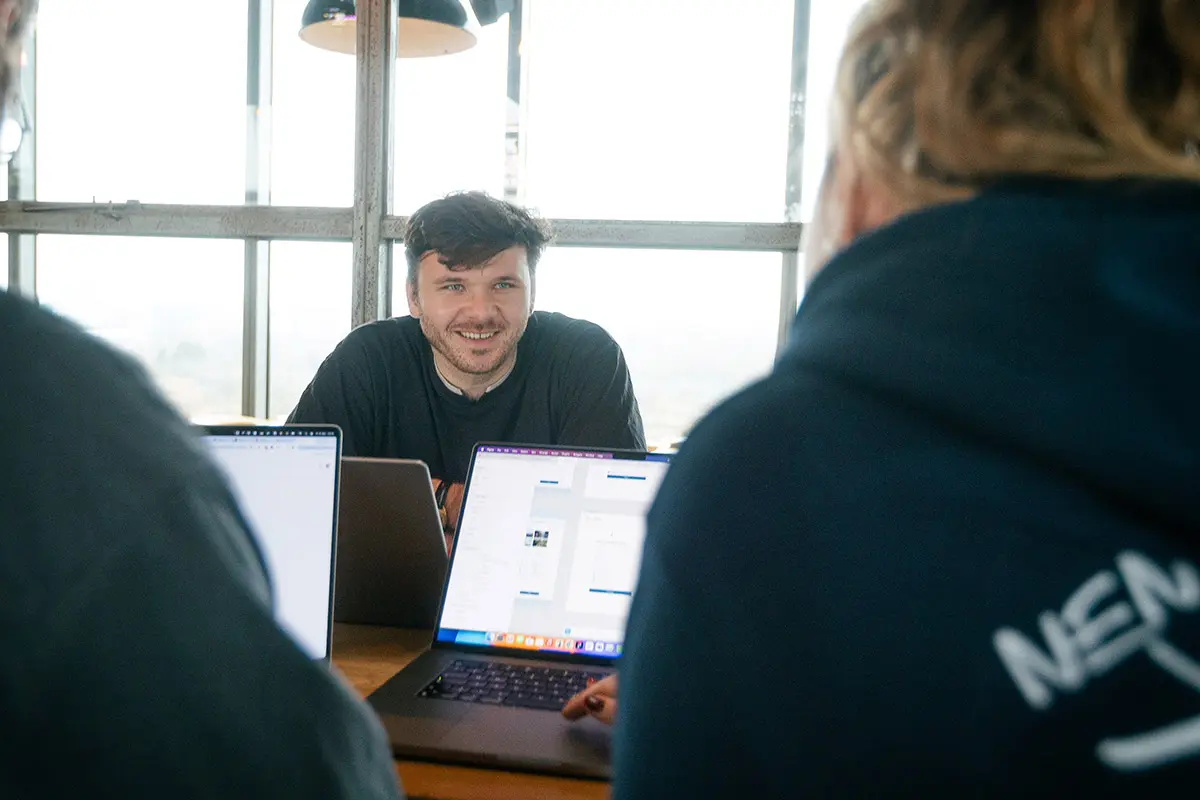 Man in a dark sweater smiling at the camera during a meeting, with laptop screens and two colleagues visible in the foreground.