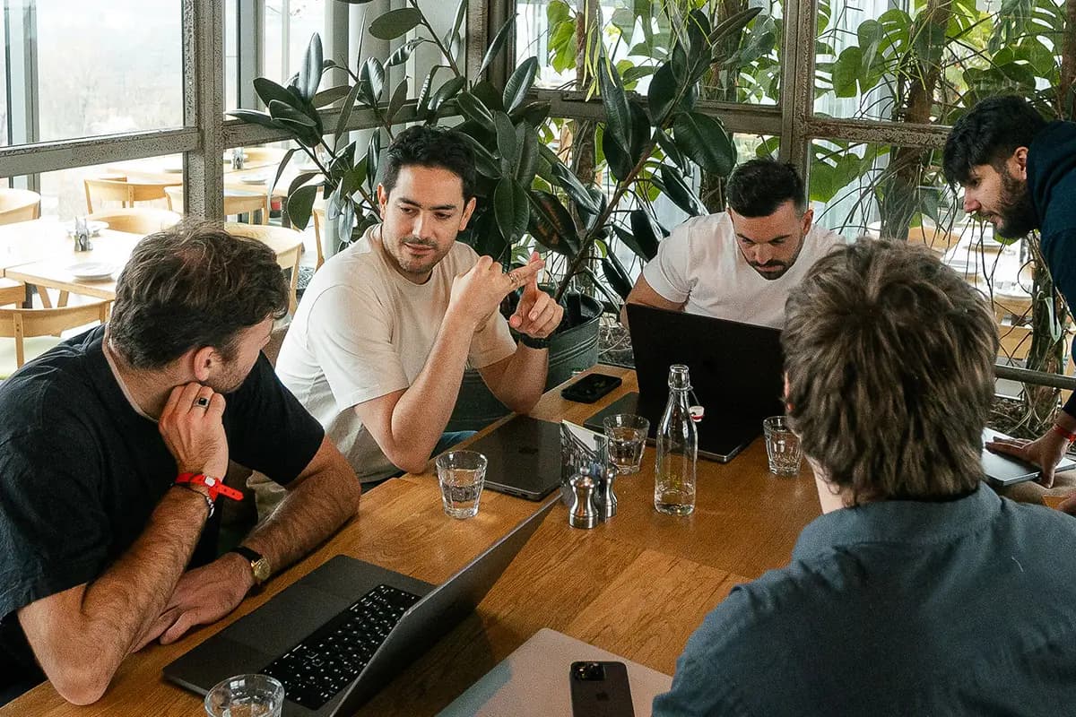Five people gathered around a table with laptops, one man gesturing mid-explanation while others listen, potted plants in the background.