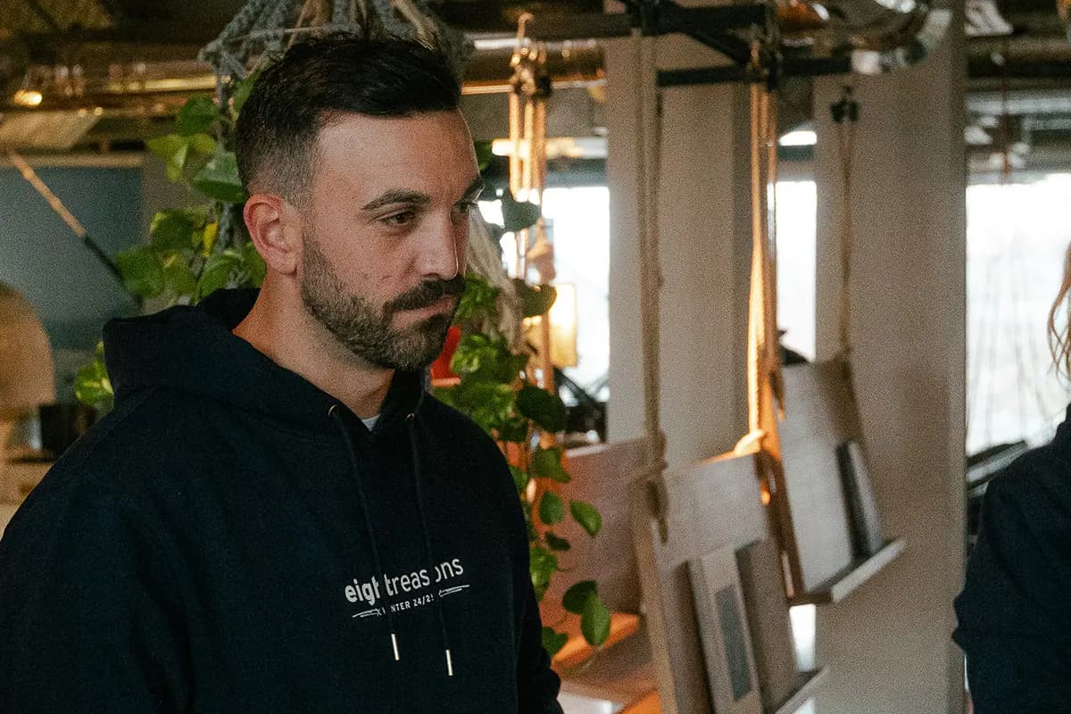Man with a beard wearing a black "eightreasons" hoodie, looking to the side in an industrial-style room with hanging plants and exposed ceiling fixtures.