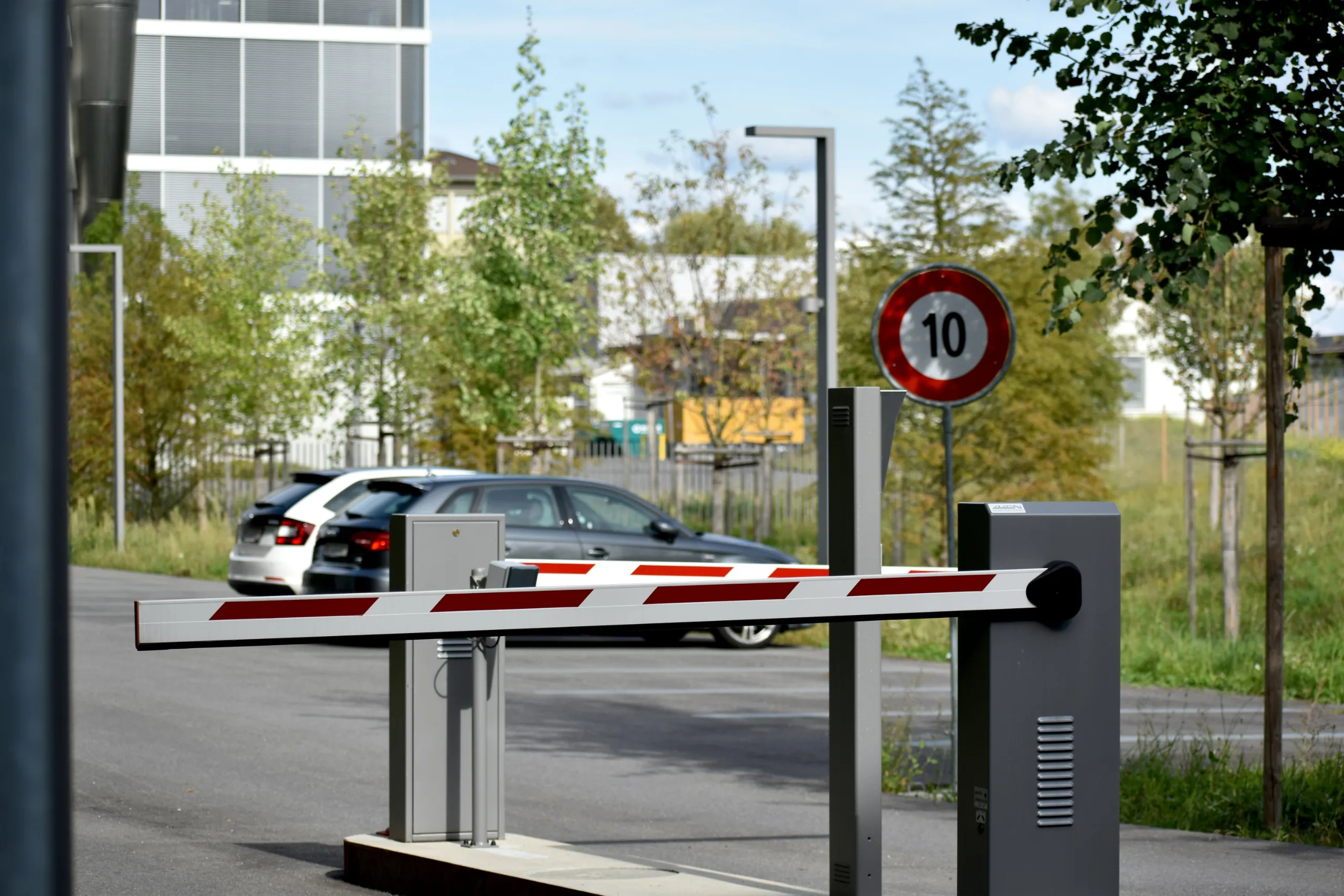 Lowered red-and-white striped vehicle barrier at a site entrance, with a 10 km/h speed limit sign mounted above and a car visible on the road behind.