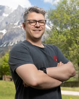 Person wearing glasses and an iits-consulting logo T-shirt standing with arms crossed, smiling, with a snow-capped mountain and green trees in the background.