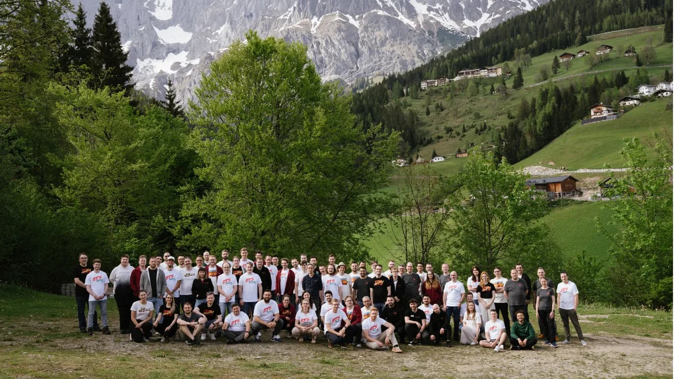 Large group of people posing for a team photo outdoors, many wearing matching white and dark branded T-shirts, with an Alpine mountain landscape, green slopes, and scattered chalets in the background.